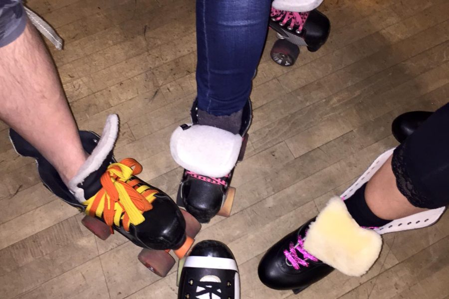 Photograph of four legs of people with roller skates on from above. Logo of SK8 Jam.