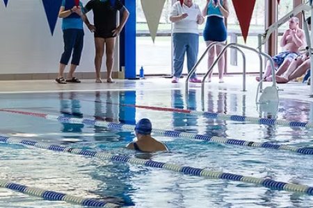 Photograph of swimming pool with people on poolside watching a swimmer. Plus Wimborne Wagtails Logo