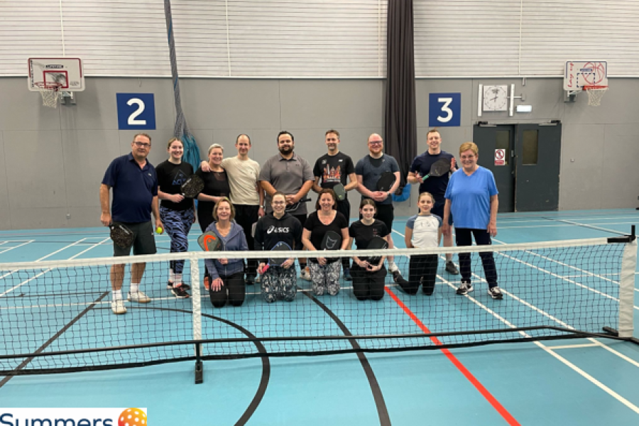 Photograph of a group of people standing behind a pickleball net and Summers Pickleball Logo