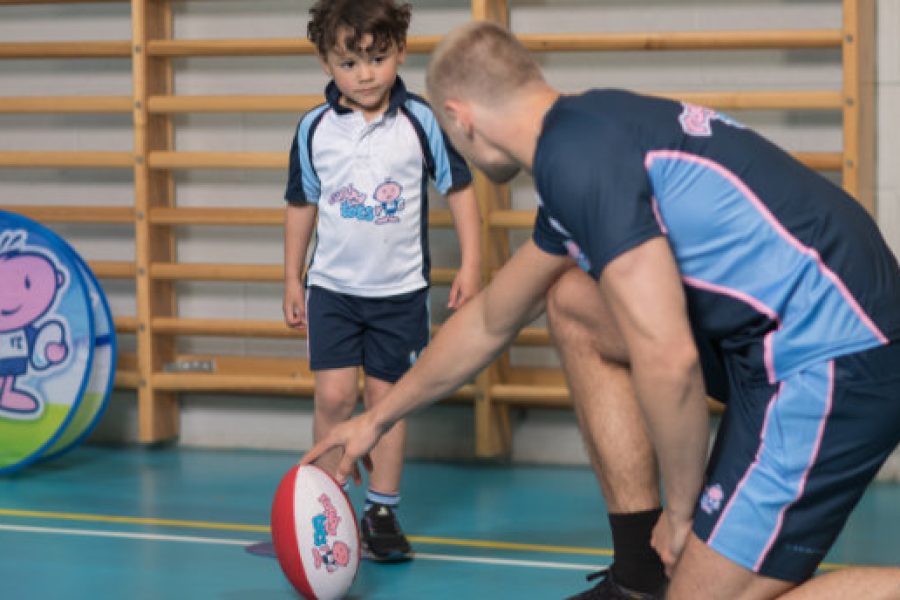 Coach in gym holding a rugby ball and talking to a young child plus logo for Rugbytots
